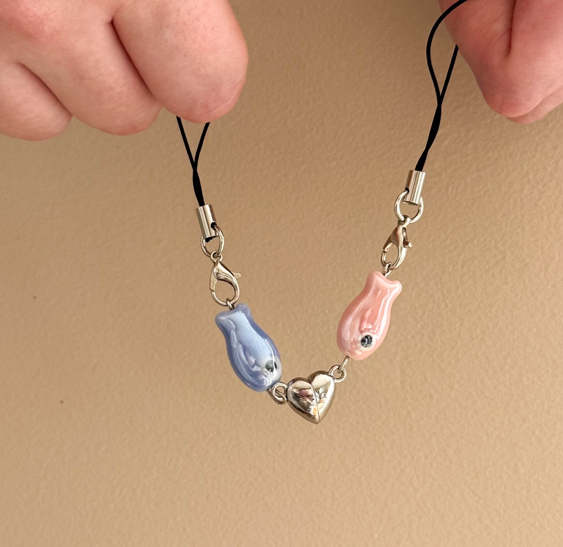 Two phone charm straps with fish and heart designs on a beige background